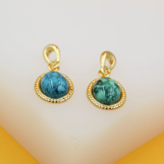 Two gold-rimmed turquoise stone charms rest on a neutral surface, displaying their detailed bead-textured settings and vibrant blue-green marbled patterns.