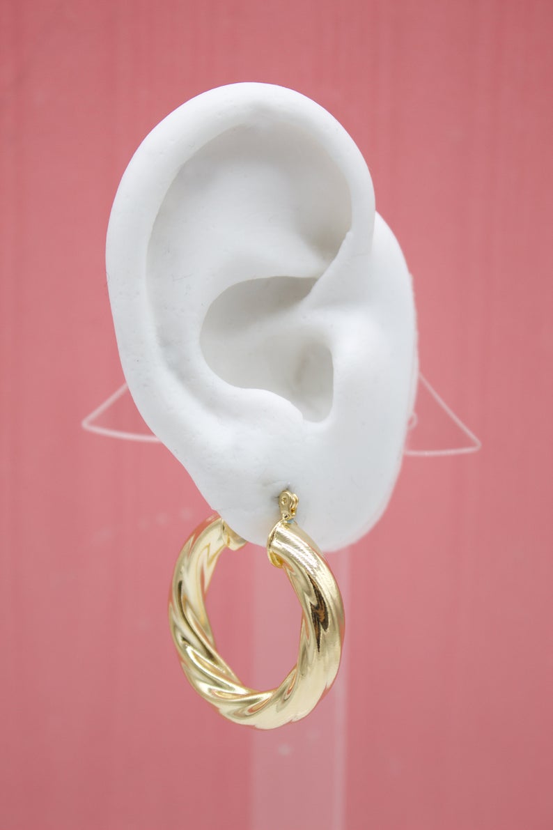 A white mannequin ear displays a shiny gold twisted hoop earring against a soft pink background.