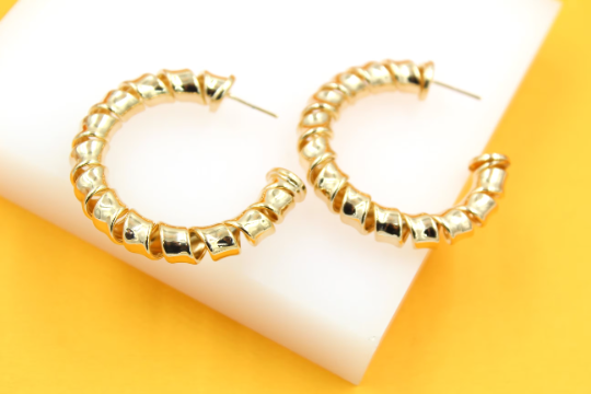 A pair of shiny gold-plated spiral hoop earrings rests on a white platform against a bright yellow background.