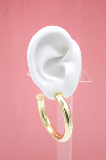 A thick gold hoop earring hangs from a white mannequin ear against a solid pink background.