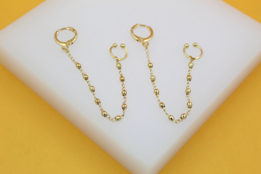 A pair of gold double piercing earrings featuring dangling bead chains resting on a white display stand.