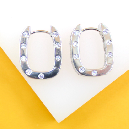 A pair of silver horseshoe-shaped hoop earrings encrusted with sparkling cubic zirconia stones rests on a white and yellow display surface.