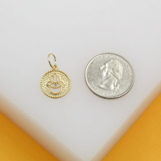 A delicate gold Hamsa hand charm with a central crystal sits next to a US quarter coin for scale on a white surface.