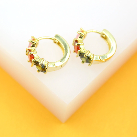 A pair of gold huggie hoop earrings adorned with colorful gemstones rests on a white and orange surface.