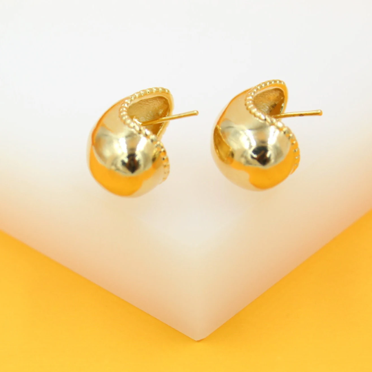 A pair of polished gold chunky huggie earrings with beaded trim rests on a white and yellow display stand.