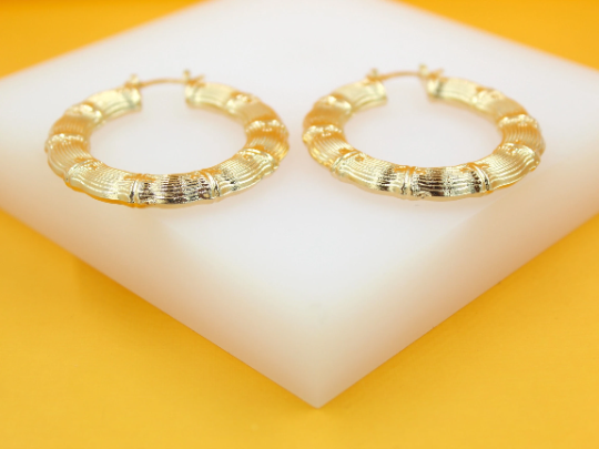A pair of glossy gold bamboo hoop earrings rests on a white display stand against a bright yellow background.