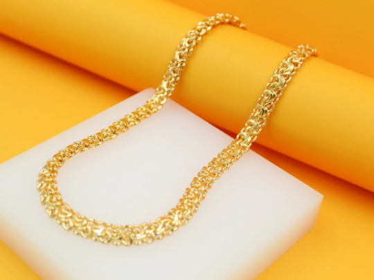 A shiny gold Byzantine chain necklace rests gracefully on a white display block against a vibrant yellow background.