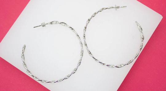 A pair of large, twisted silver hoop earrings rests on a white display stand against a vibrant pink background.