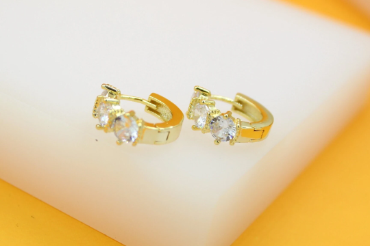 A pair of gold huggie hoop earrings embedded with sparkling cubic zirconia stones rests elegantly on a white surface.