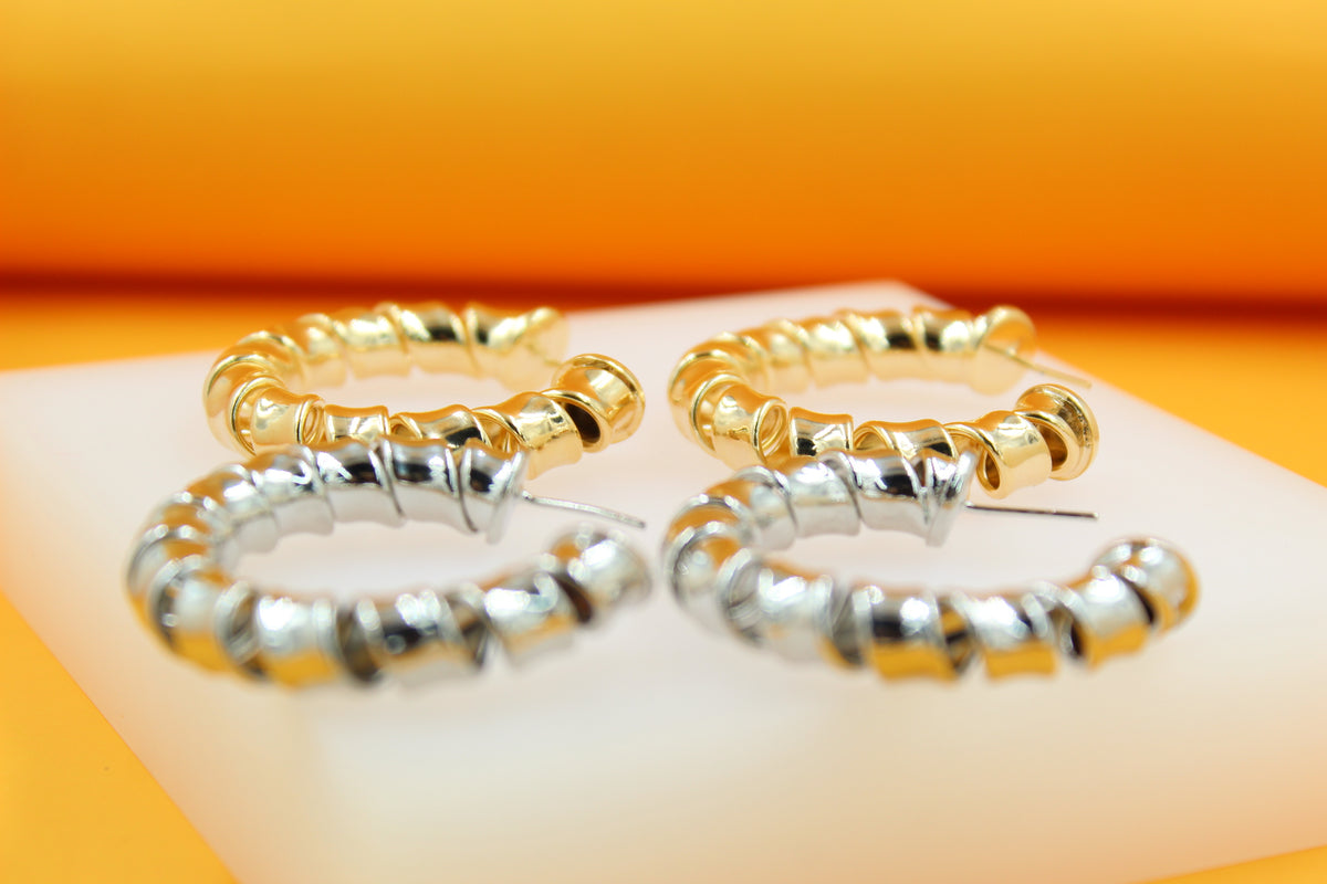 Two pairs of chunky, spiral-textured hoop earrings—one pair in gold and the other in silver—rest on a white platform against a warm orange background.