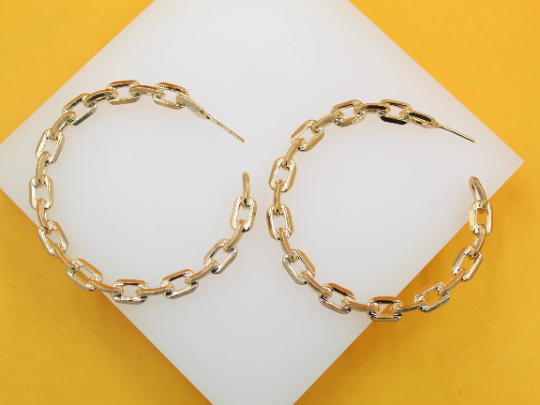 A pair of shiny gold chain link hoop earrings rests on a white display surface against a vibrant yellow background.