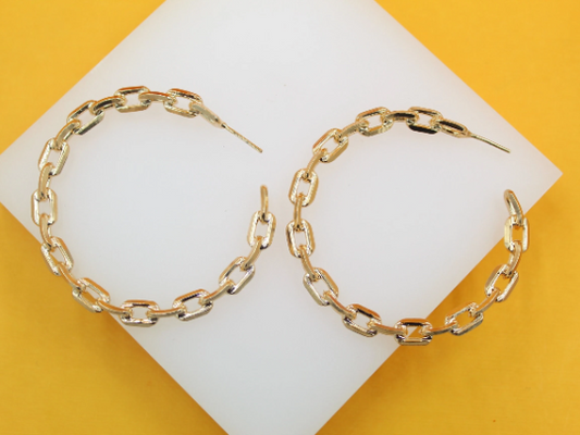 A pair of shiny gold chain link hoop earrings rests on a white display surface against a vibrant yellow background.