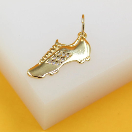 A gold soccer cleat charm rests on a white display block, featuring sparkling cubic zirconia stones along the side stripes.