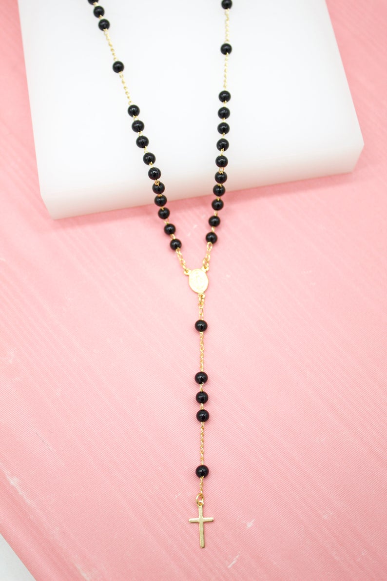 A delicate gold rosary necklace featuring glossy black beads and a small crucifix rests against a soft pink and white background.