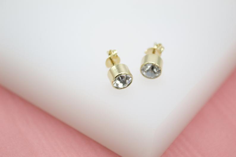 A pair of gold bezel-set crystal stud earrings rest on a clean white display surface.