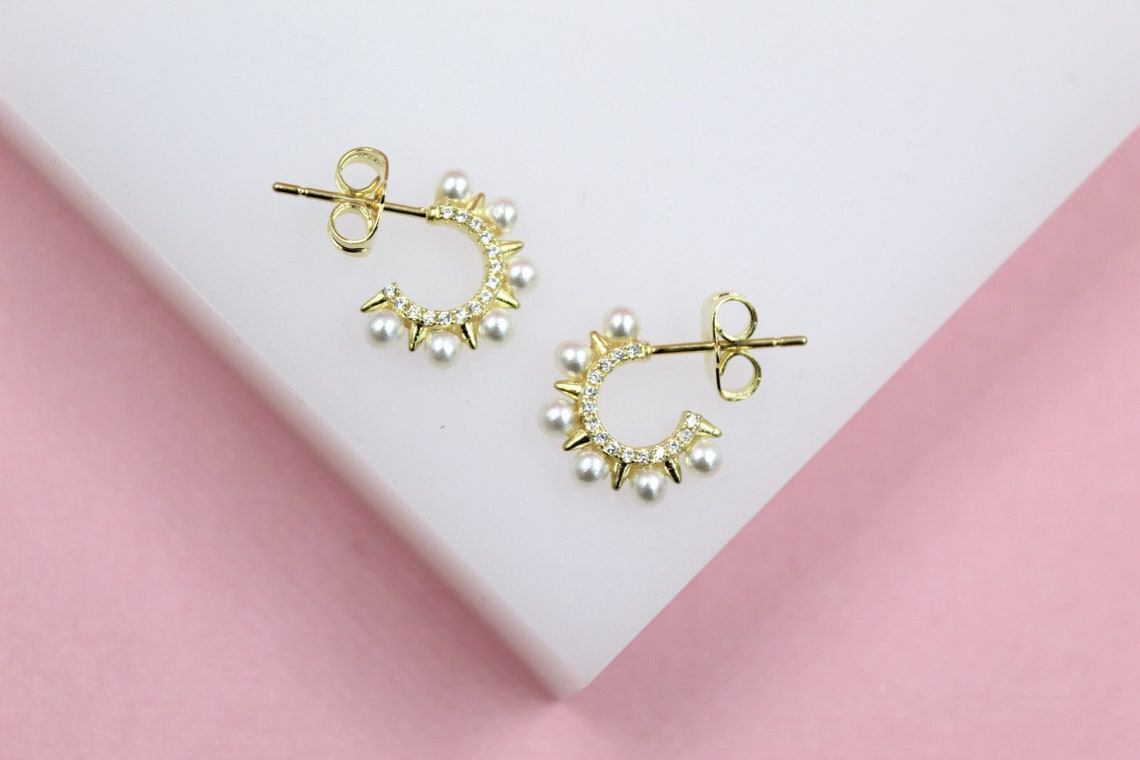 A pair of gold huggie hoop earrings featuring sparkling pave crystals and delicate pearl accents rests on a white and pink surface.