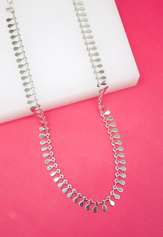 A delicate silver chain necklace features dangling teardrop-shaped charms draped across a white display block against a vibrant pink background.