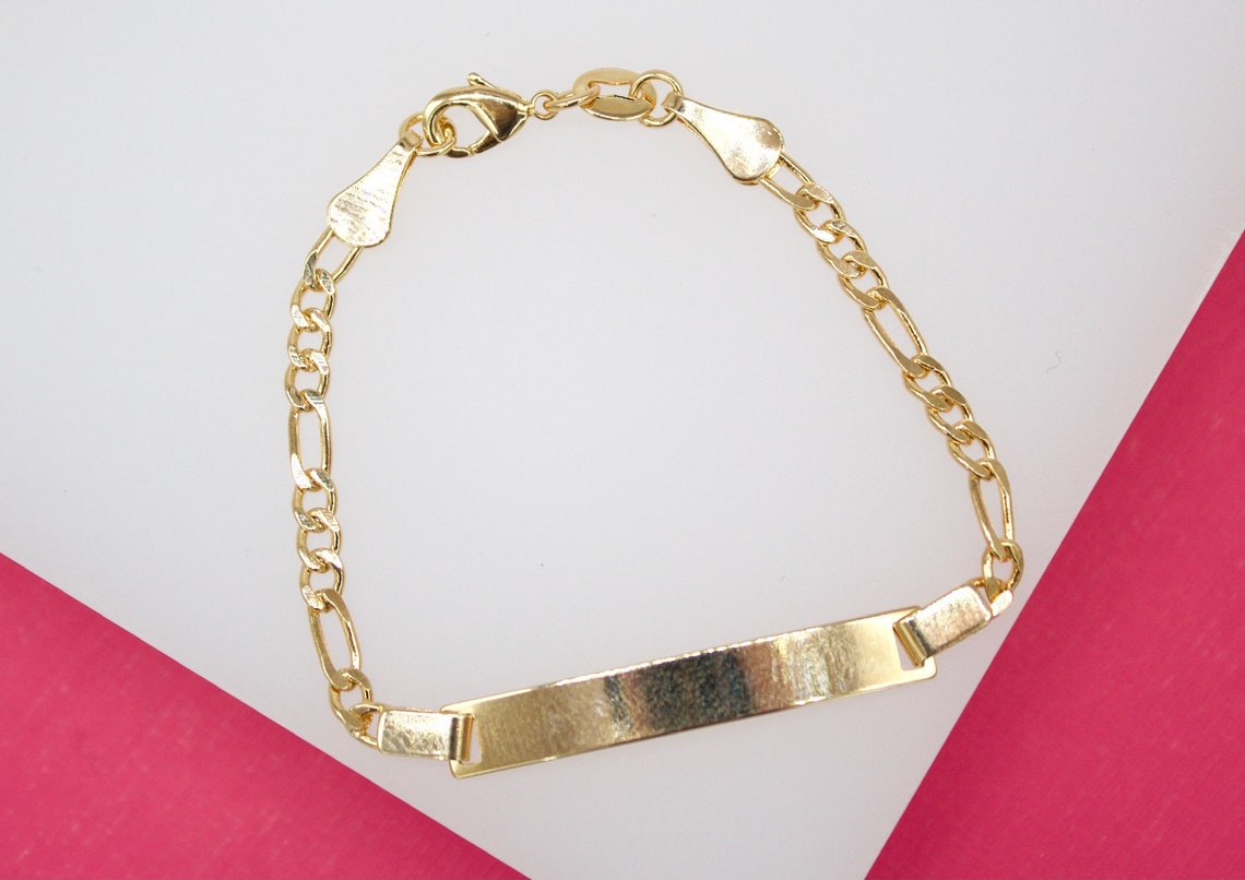 A shiny gold identification bracelet with a Figaro chain rests against a two-tone pink and white background.
