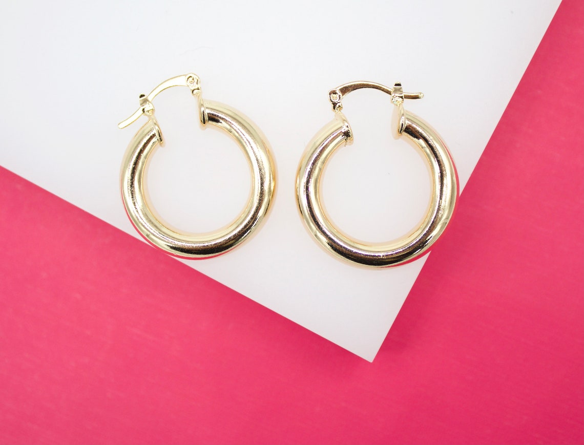Two polished thick gold hoop earrings rest against a white surface set against a vibrant pink background.
