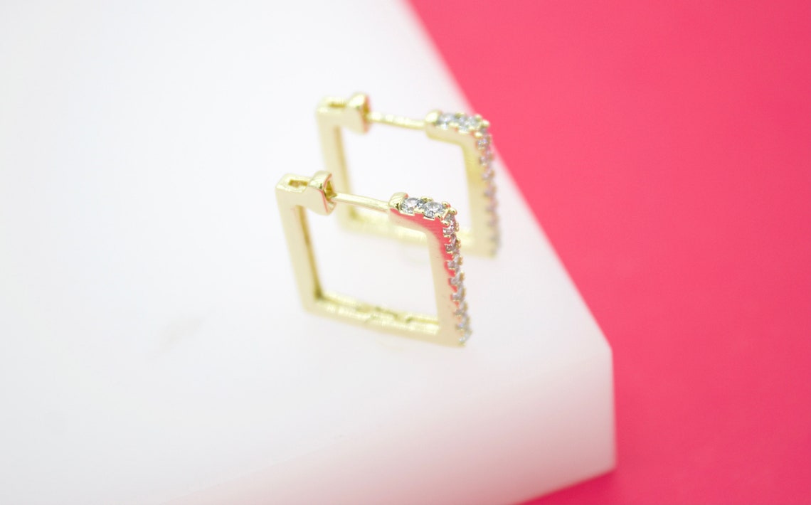 Two gold square hoop earrings embellished with sparkling CZ stones rest on a white display stand against a vibrant pink background.