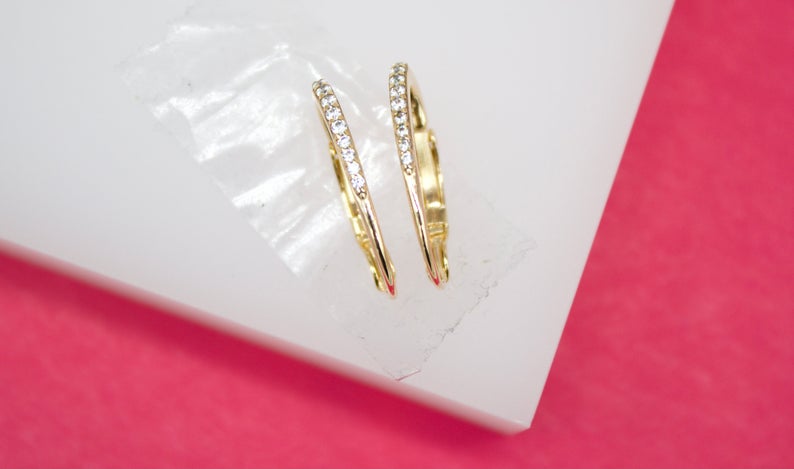 Two elegant gold hoop earrings accented with sparkling cubic zirconia crystals rest on a white display against a vibrant pink background.