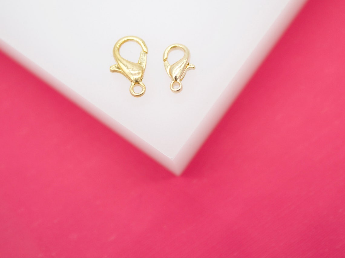 Two shiny gold lobster claw clasps rest on a white surface against a bright pink background.