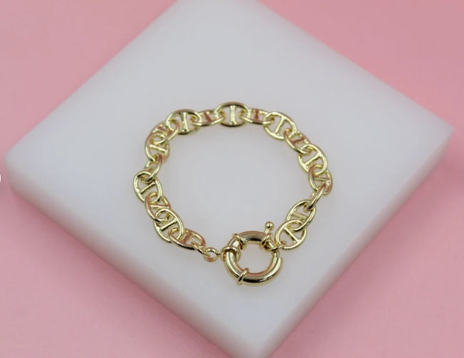 A gleaming gold anchor chain bracelet rests gracefully on a white display block against a soft pink background.