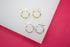 Two pairs of twisted hoop earrings, one in gold and one in silver, rest on a white display surface against a vibrant pink background.