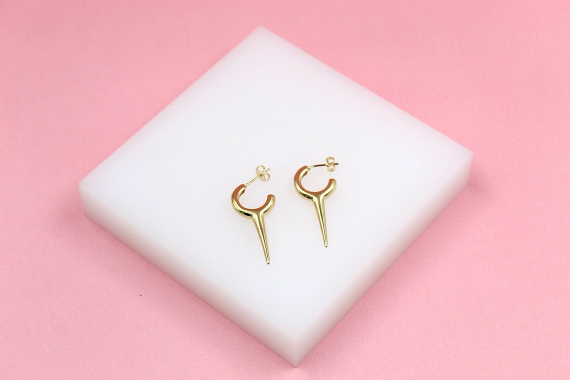 A pair of stylish gold spike hoop earrings rests on a white square display block against a vibrant pink background.