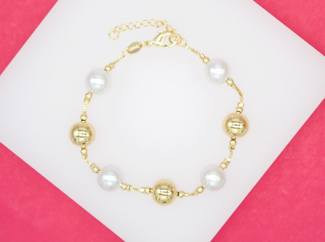 A delicate gold chain bracelet features alternating white pearls and polished gold beads on a white and pink background.