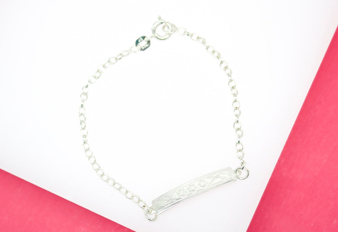 A delicate sterling silver ID bracelet rests on a white and pink background, featuring a thin link chain and an engraved rectangular plate.