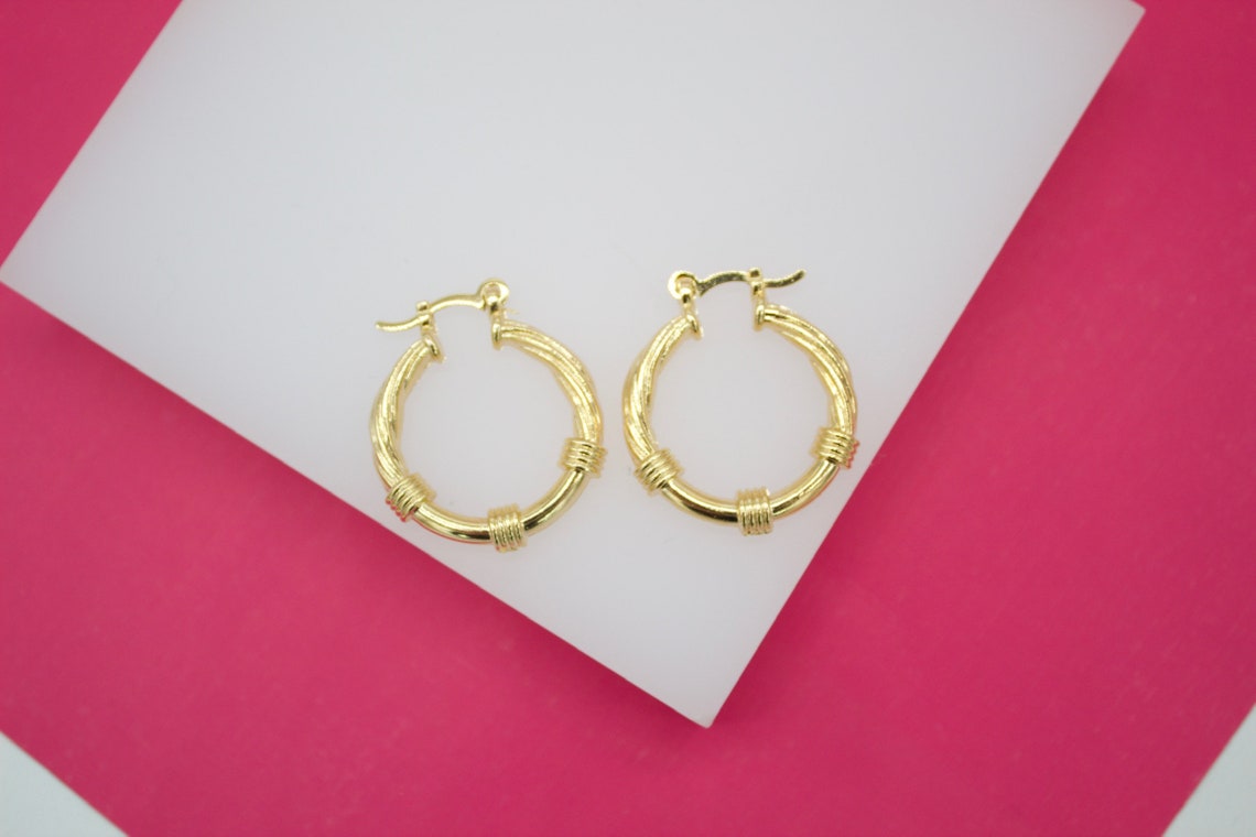 A pair of textured gold hoop earrings rests against a white display block over a vibrant pink background.
