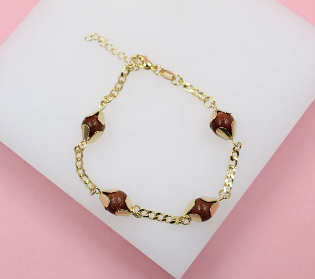 A gold-plated link bracelet features four sparkling brown sandstone bead charms cradled in delicate gold caps against a white and pink background.