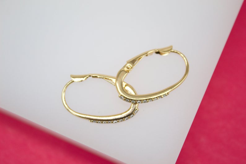 A pair of elegant gold hoop earrings with sparkling crystal accents rests on a white display surface against a pink background.