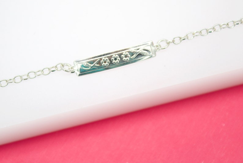 A delicate sterling silver ID bracelet rests on a white and pink display surface, showcasing an engraved floral pattern on its rectangular bar.