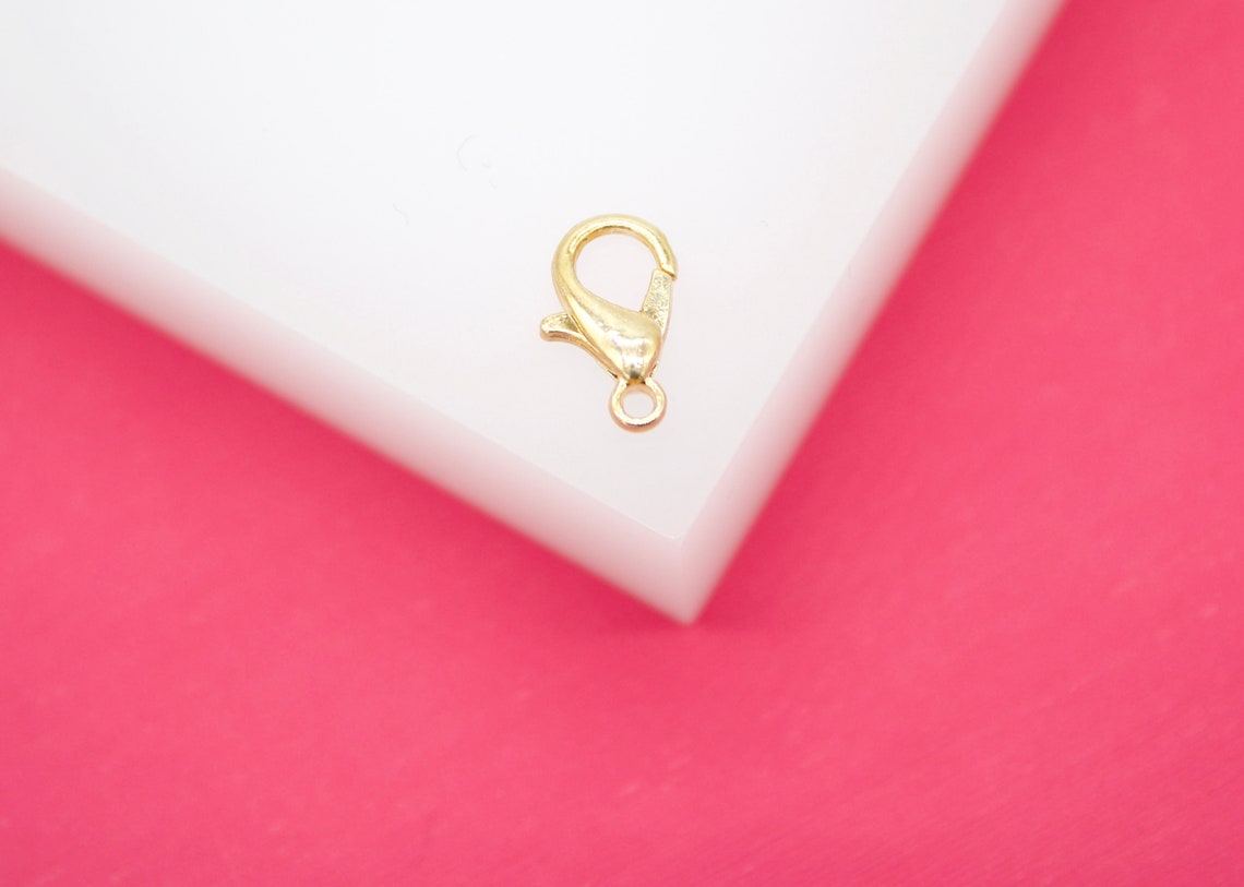 A small gold lobster claw clasp rests on a white display block against a vibrant pink background.