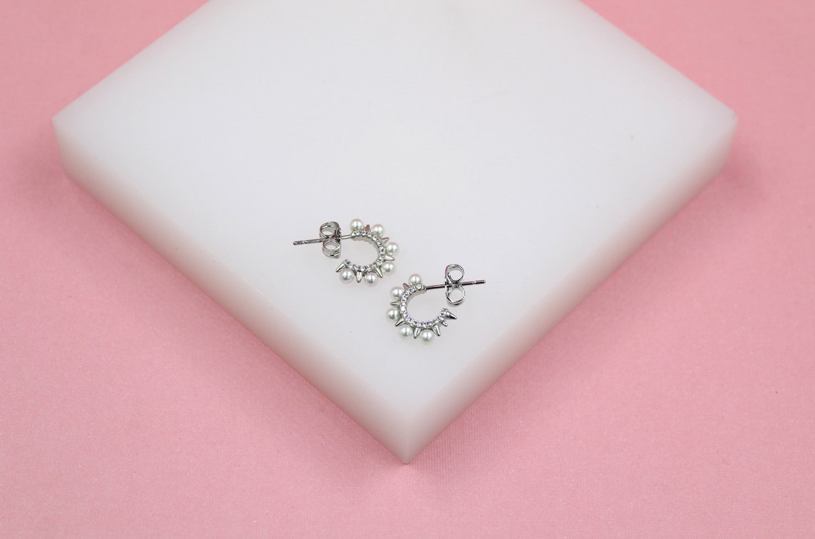 A pair of silver hoop earrings adorned with pearls and spikes rests on a white block against a pink background.