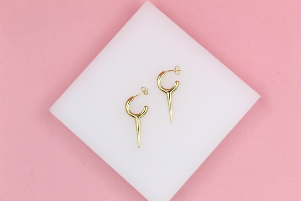 A pair of gold hoop earrings with sharp spike charms rests on a white square pedestal against a pink background.