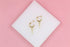 A pair of gold hoop earrings with sharp spike charms rests on a white square pedestal against a pink background.