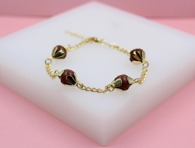 A gold-plated charm bracelet featuring four round brown sandstone beads resting on a white display block against a pink background.