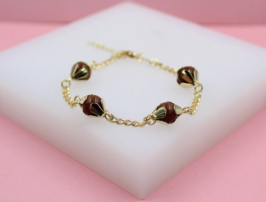 A gold-plated charm bracelet featuring four round brown sandstone beads resting on a white display block against a pink background.