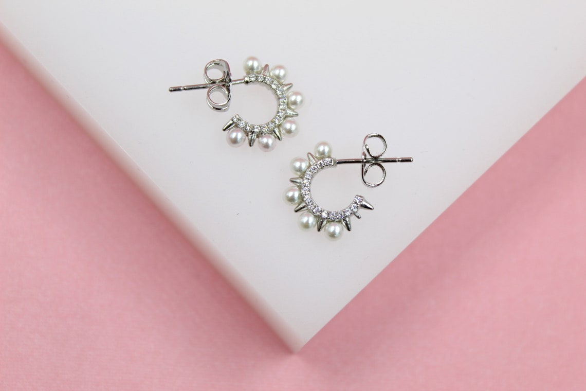 A pair of silver hoop earrings adorned with tiny pearls and sparkling crystals rests on a white display against a pink background.
