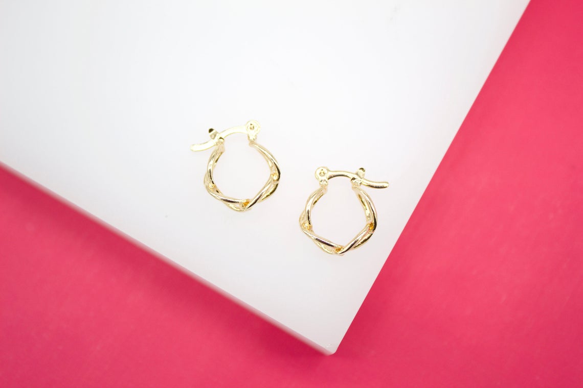 A pair of small, twisted gold hoop earrings rests on a white display against a vibrant pink background.
