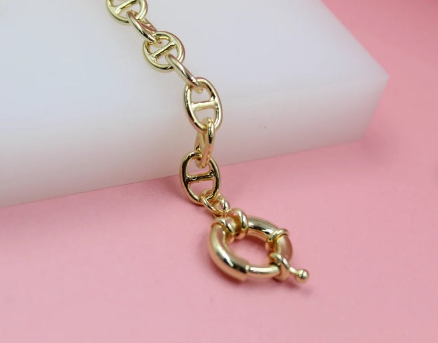 A shiny gold mariner chain rests on a white and pink background, highlighting its secure round clasp.