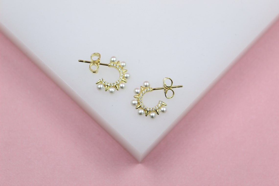 A pair of gold huggie hoop earrings featuring pearl accents and sparkling cubic zirconia rests on a white display surface against a pink background.
