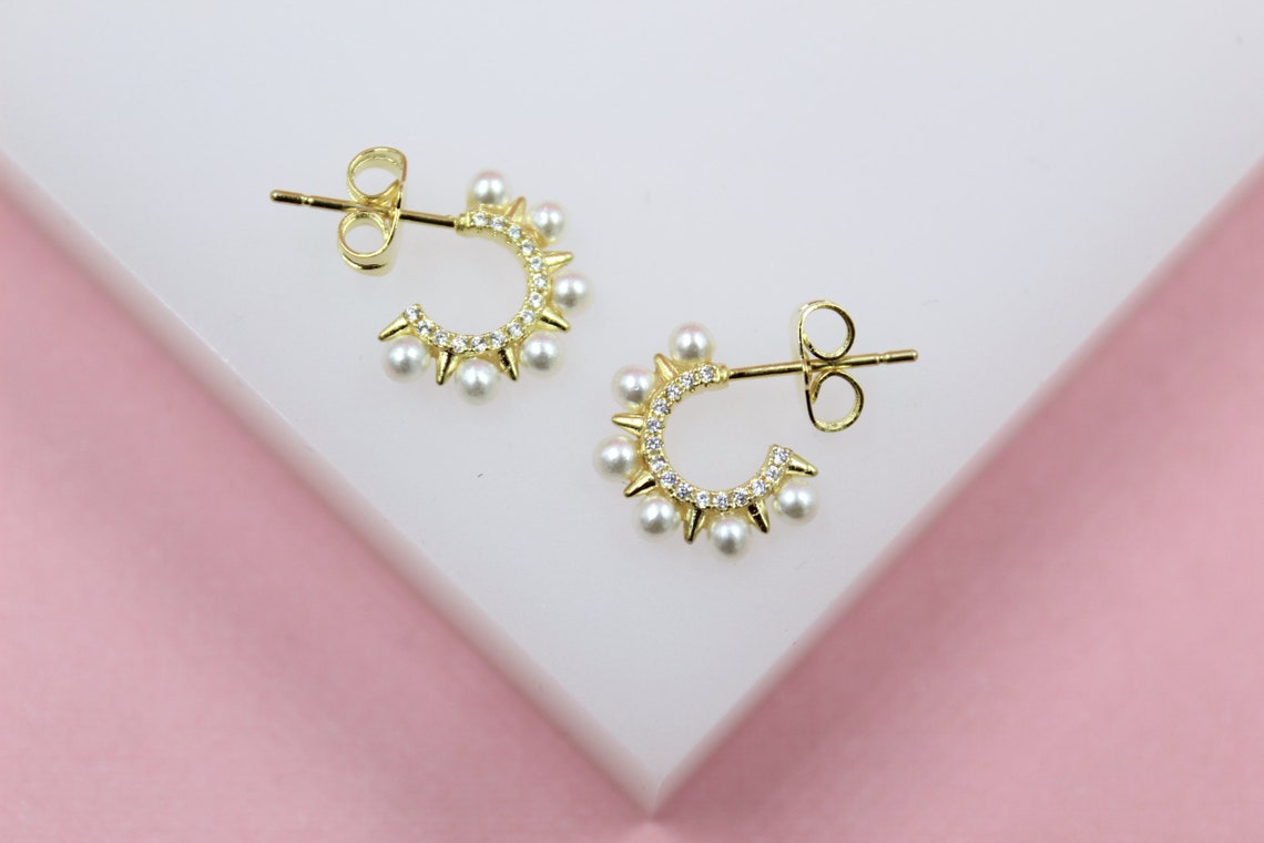 Two gold hoop earrings featuring delicate pearls and sparkling pave stones rest on a white surface against a pink background.