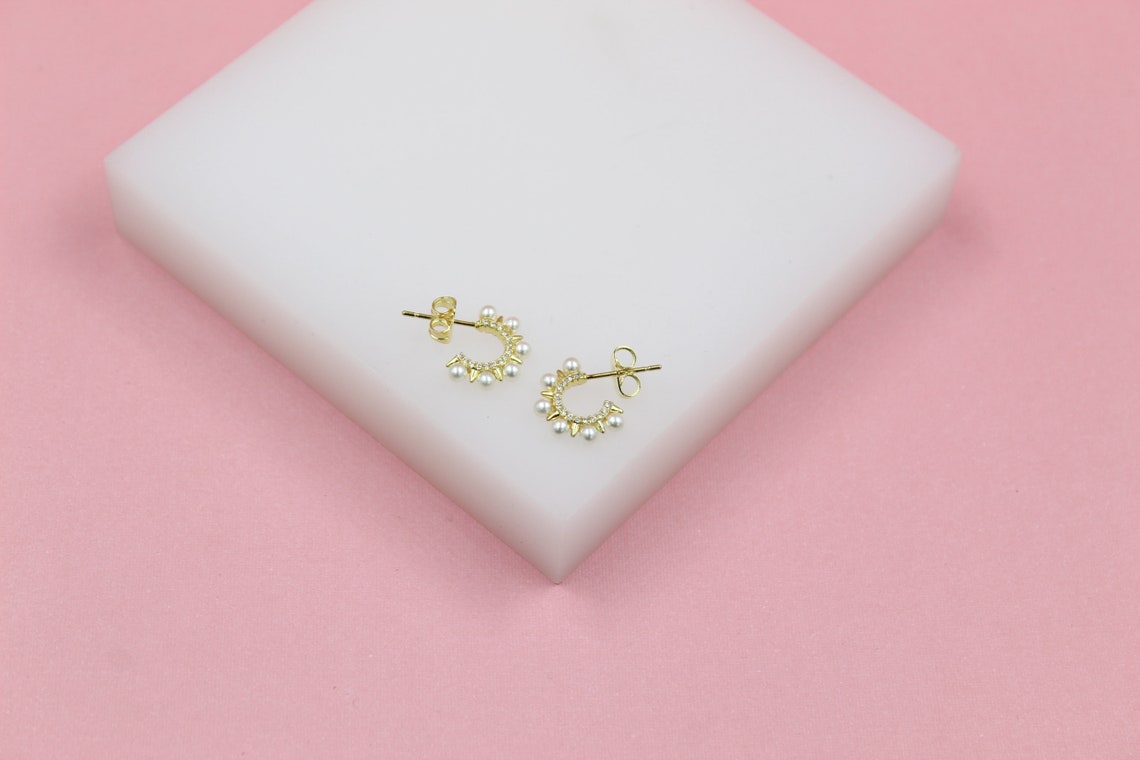 A pair of gold hoop earrings embellished with pearls and tiny spikes rests on a white display block against a pink background.