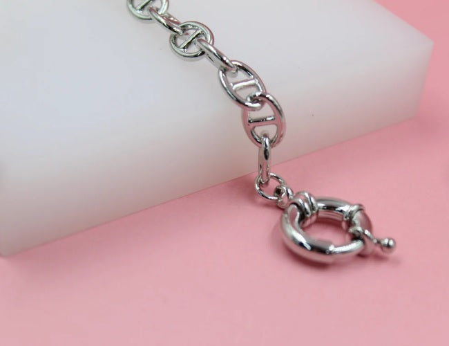 A shiny silver chain bracelet featuring distinctive anchor links and a secure round clasp rests on a white and pink display surface.