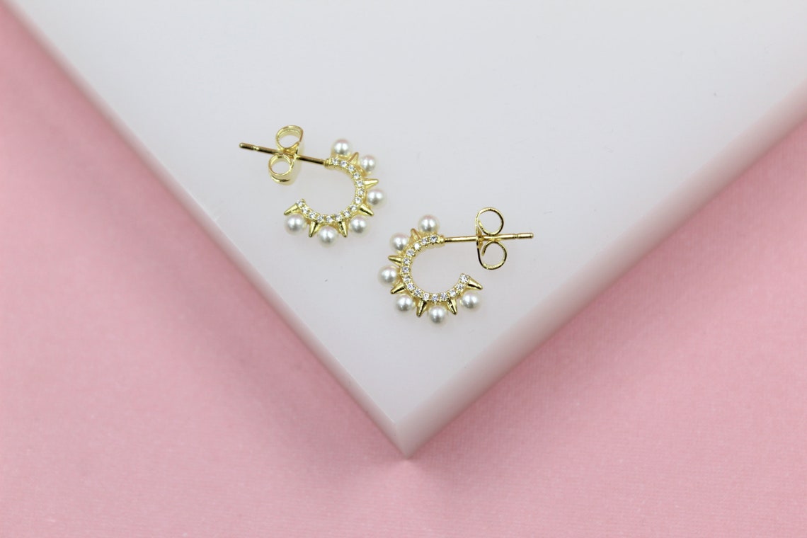 A pair of gold huggie hoop earrings decorated with tiny pearls and sparkling crystals rests on a white display against a pink background.