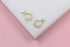 A pair of gold huggie hoop earrings decorated with tiny pearls and sparkling crystals rests on a white display against a pink background.